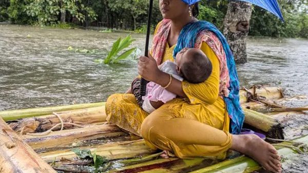 a woman sitting on a bamboo raft during a flood, holding a baby under an umbrella in Bangladesh Climate Crisis