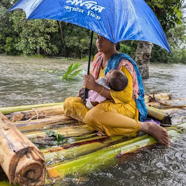 a woman sitting on a bamboo raft during a flood, holding a baby under an umbrella in Bangladesh Climate Crisis